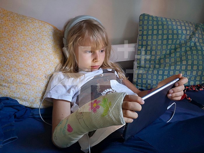 8 year old girl with broken, plastered wrist, watching a cartoon on a touch pad, European, France.