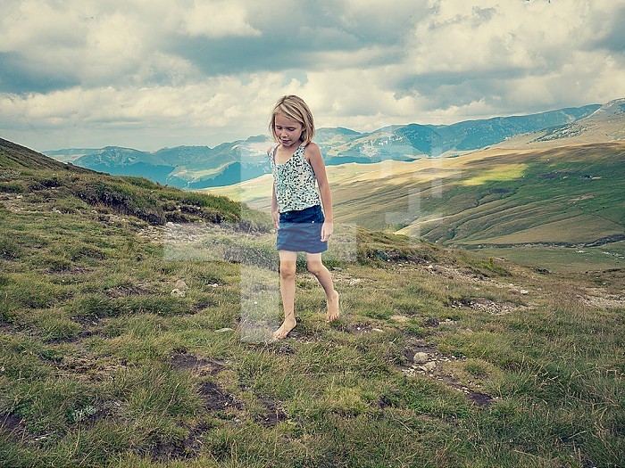 Girl 6-9 years old, European type with blond hair walking facing front looking down at the ground barefoot on a clear cloudy day in the mountains in the Carpathians in the summer, Romania, Europe