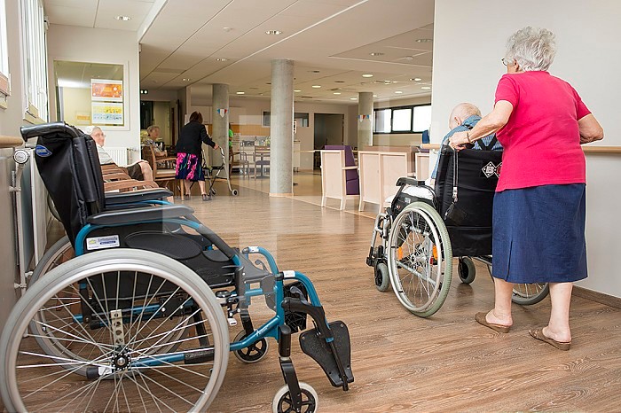 EHPAD - Elderly woman pushing her husband, one of the residents, in a wheelchair. Empty wheelchair and boarders in a common room.