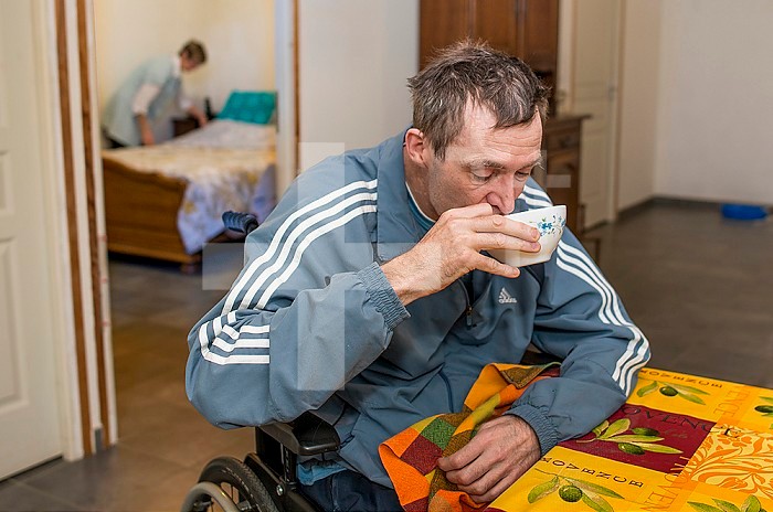 ADMR 62 - Home Help in Rural Areas, Pas de Calais. Muriel, Auxiliary of Social Life (AVS) works at the home of Mr. T. who is severely handicapped by Multiple Sclerosis. Muriel must help her with all the daily tasks. Disabled person in his wheelchair having breakfast and his carer making the bed in the bedroom.