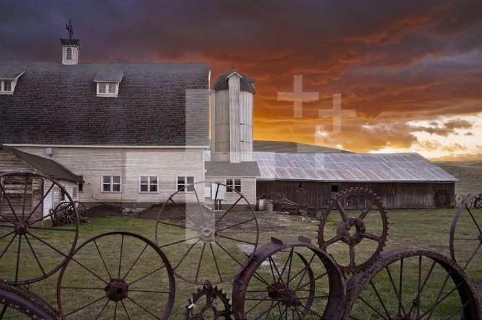 A fence of wagonwheels and large gears in front of a large barn at dusk, Washington, USA.